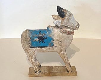 Puede incluir: Una figura de toro tallada en madera a mano con un acabado blanquecino desgastado, cuernos negros y una manta de silla de montar azul brillante con un patrón floral negro. El animal se apoya sobre una base rectangular y lleva un collar decorativo.