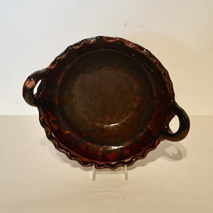 Op de afbeelding: Een bruine keramische kom met twee handvatten. De kom heeft een gegolfde rand en een geglazuurde afwerking.