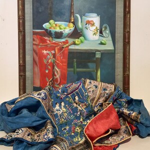 May include: A framed painting of a still life with fruit, a teapot, and a musical instrument. The painting is in a wooden frame with a bamboo-like design. A blue and red silk fabric with embroidered figures is in the foreground.