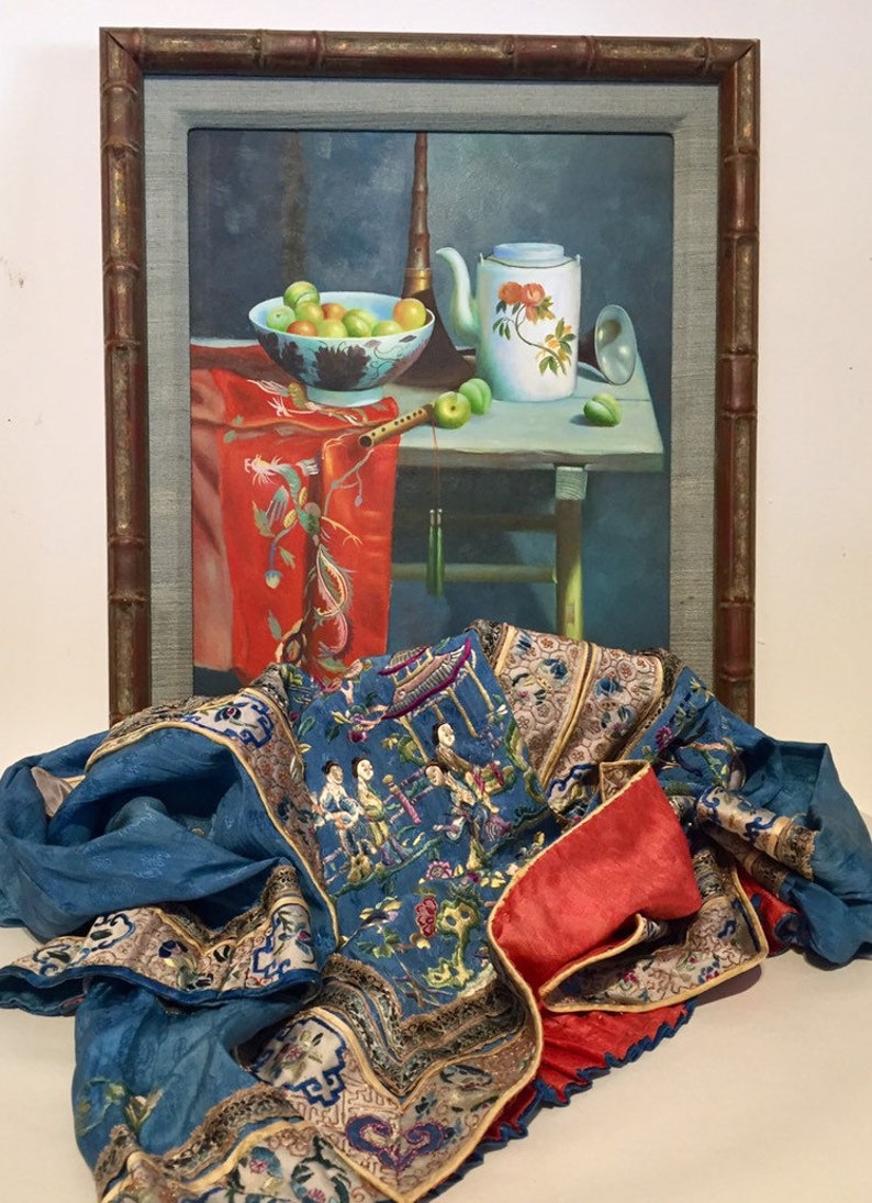 May include: A framed painting of a still life with fruit, a teapot, and a musical instrument. The painting is in a wooden frame with a bamboo-like design. A blue and red silk fabric with embroidered figures is in the foreground.