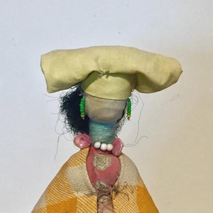 May include: A handmade doll with a yellow fabric hat, black hair, and a pink and white checkered dress. The doll is wearing a necklace of white beads and green beaded earrings.