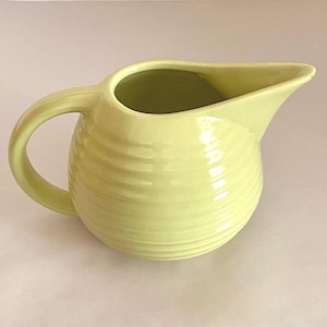 May include: A pale yellow ceramic pitcher with a rounded body and a ribbed design. The pitcher has a handle and a spout.