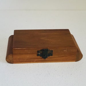 May include: A small, rectangular wooden box with a hinged lid and a dark metal clasp. The box is made of light brown wood with rounded edges and a slightly raised top. The clasp is centered on the front of the box.