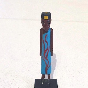 May include: A wooden figurine of a person wearing a blue and red robe and a black hat with a yellow design. The figurine is standing on a black base.