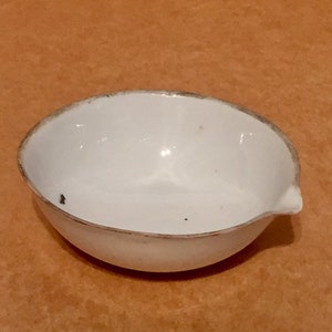 Puede incluir: Un crisol de porcelana blanca con un pequeño pico. El crisol está ligeramente descolorido alrededor del borde.