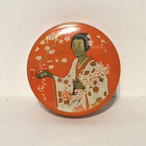 May include: A round orange tin with a white and gold design of a woman in a kimono holding a bouquet of flowers. The woman is surrounded by cherry blossoms.