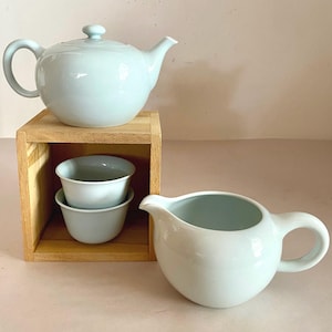 Op de afbeelding: Een lichtblauwe theepot, roomkan en twee theekopjes van keramiek. De theepot en roomkan hebben een afgeronde vorm. De theekopjes zijn gestapeld in een houten doos.