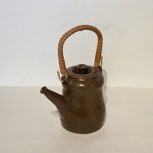 May include: A brown ceramic teapot with a woven wicker handle and lid. The teapot has a rounded body and a spout.