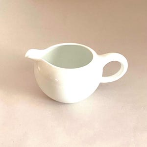 Op de afbeelding: Een witte keramische roomkan met een rond lichaam en een enkele handgreep. De kan is ontworpen om vloeistoffen zoals room of melk te gieten.