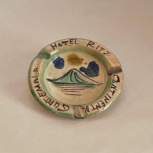 Puede incluir: Un cenicero de cerámica con un diseño pintado en verde, azul y amarillo de una montaña y nubes. El cenicero está inscrito con el texto "Hotel Ritz Guatemala" y "Continent Bl".