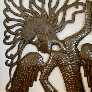 May include: A metal wall art piece depicting an angel with outstretched arms. The angel has detailed wings and a sun-like halo. The artwork is made of a dark, metallic material and is mounted on a white wall.