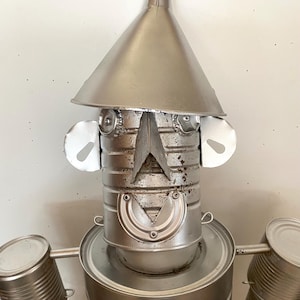 Peut inclure: Sculpture métallique artisanale représentant un robot. La tête est un entonnoir, avec des yeux en capsules de bouteilles et des oreilles en boîtes de conserve. Le corps est fait de boîtes recyclées, avec des bras et un emblème en forme de cœur. Esthétique industrielle.