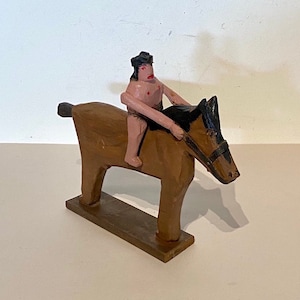 May include: A wooden figurine of a person riding a horse. The person is painted pink with black hair and a black hat. The horse is brown with a black mane and bridle. The figurine is on a wooden base.