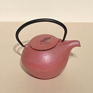 May include: A pink cast iron teapot with a black handle and lid. The teapot has a textured surface.
