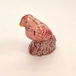 May include: A small pink bird figurine with a yellow beak, perched on a textured, red base. The bird has a distressed paint finish, giving it a vintage look.