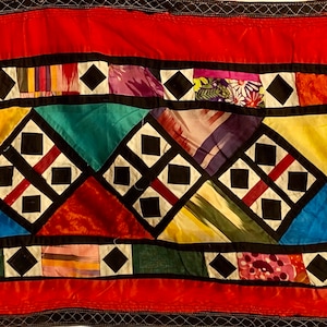 以下が含まれることがあります： 黒色の正方形と三角形が幾何学模様になった、カラフルなパッチワークキルト。赤、黄、青、緑、白の様々な生地で作られています。