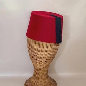 Peut inclure: Un fez rouge vif doté d'un sommet plat et d'un long pompon bleu foncé attaché au centre. Le chapeau présente une forme cylindrique avec une texture lisse semblable à du feutre et est présenté sur un support de tête en osier tressé.