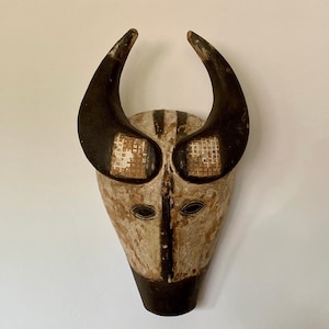 Può includere: Una maschera di legno con un designo dipinto in bianco e nero. La maschera ha due grandi corna che si curvano verso l'alto. La maschera ha un naso lungo e appuntito e due piccoli occhi.