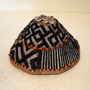 Puede incluir: Sombrero de cuentas con forma cónica, con un patrón geométrico en blanco y negro. El sombrero está adornado con cuentas rojas y amarillas y adornado con conchas marinas en la parte superior. La base es de color beige.