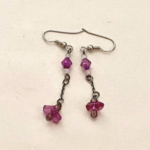 May include: A pair of dangle earrings with silver-tone hooks. Each earring features a series of small, clear beads, a faceted purple bead, and a cluster of pink flower-shaped beads. The earrings are connected by a delicate chain.