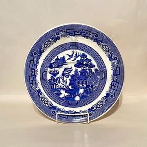 May include: A blue and white Willow pattern plate. The plate features a detailed landscape scene with a bridge, buildings, and trees. The rim has a decorative border. The plate is displayed on a white metal stand.