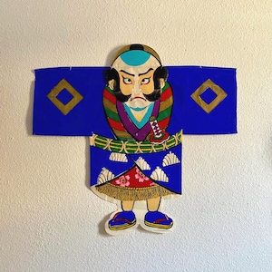 May include: A colorful, stylized Japanese Kabuki actor figure. The figure is wearing a blue kimono with gold diamond accents, a multi-colored top, and a red and white skirt. The actor has a stern expression and is holding a sword.