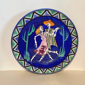 Puede incluir: Un plato de cerámica azul con un borde decorativo de triángulos verdes y blancos. El centro del plato presenta una escena pintada de dos esqueletos bailando, uno con un vestido rosa y el otro con una camisa a rayas verdes y blancas y un sombrero amarillo. La escena se desarrolla sobre un fondo azul con cactus verdes.