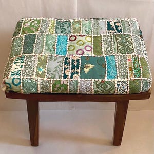 Könnte beinhalten: Ein kleiner Holzhocker mit einem Patchwork-Kissen. Das Kissen ist aus verschiedenen Stoffen in Grün- und Blautönen mit Stickereien gefertigt. Der Hocker hat zwei Holzbeine.