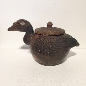 May include: A brown ceramic duck-shaped container with a lid. The container has a textured surface and is designed with carved details. The lid has a small knob on top. The duck is positioned on a white surface.