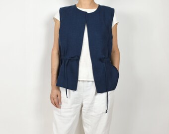 May include: A navy blue linen vest with a drawstring closure. The vest has a relaxed fit and is open in the front. The model is wearing the vest over a white shirt and white pants.
