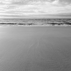 May include: A black and white landscape photograph featuring a wide, sandy beach in the foreground with gentle waves rolling onto the shore. In the distance, the horizon line meets a cloudy sky, with faint silhouettes of wind turbines visible.