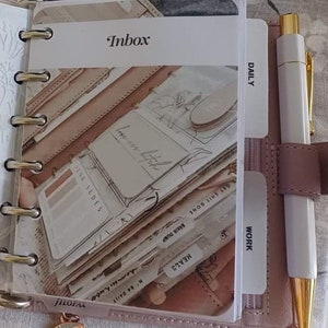 Puede incluir: Una agenda de cuero rosa con bolsillos de plástico transparente y pestañas. Las pestañas están etiquetadas como "Inbox", "Daily", "Work", "Meals", "Brain Dump" y "Get It Done". Un bolígrafo blanco con una tapa dorada está en la agenda.