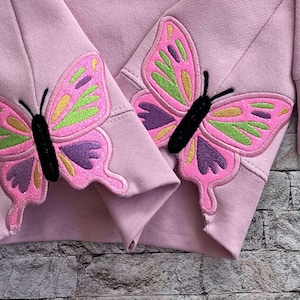 Puede incluir: Una sudadera rosa con diseños de mariposas bordadas en las mangas. Las mariposas son rosas, verdes y moradas.