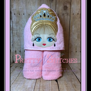 Può includere: Asciugamano con cappuccio rosa con un viso di ragazza ricamato con una corona sulla testa. L'asciugamano è piegato e in piedi su una superficie di legno. Il testo "Pretty Stitches" è ricamato sull'asciugamano.