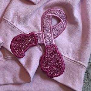 May include: Pink sweatshirt with a pink glitter embroidered patch of a pink ribbon with boxing gloves on either side.
