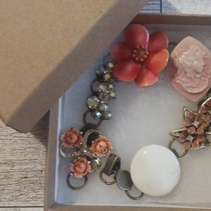 Puede incluir: Un brazalete vintage con un dije de flor rosa, un dije de cerámica blanca y un grupo de dijes de flores rosas más pequeños. El brazalete está hecho de metal dorado y tiene un diseño delicado y femenino.