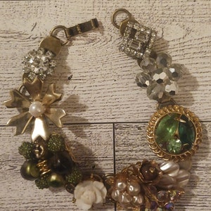 Puede incluir: Un brazalete de tono dorado con una variedad de encantos, incluyendo una flor, una gema verde y un racimo de perlas. El brazalete está hecho de metal y tiene un cierre de broche.