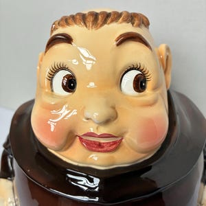 May include: A ceramic cookie jar in the shape of a jovial monk's head. The jar features a detailed face with rosy cheeks, brown eyes, and a smiling red mouth. The monk wears a brown robe and has a bald head with a brown hair outline.