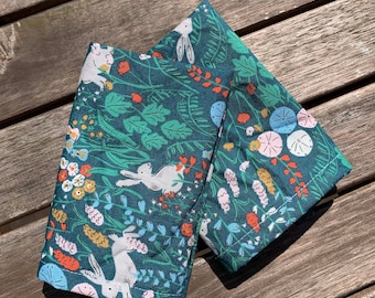 May include: Two green fabric napkins with a white bunny rabbit and floral pattern. The napkins are folded in half and have a white border.