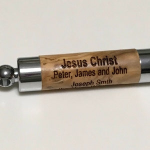 Peut inclure: Un bouchon de bouteille cylindrique avec une section centrale en bois et des extrémités en métal poli. La section en bois présente le texte gravé : "Jesus Christ, Peter, James and John, Joseph Smith."