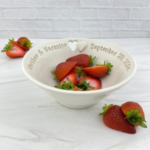 May include: White ceramic bowl with a heart-shaped cutout and the inscription "Matthew & Veronica September 28, 2021". The bowl is filled with fresh strawberries.