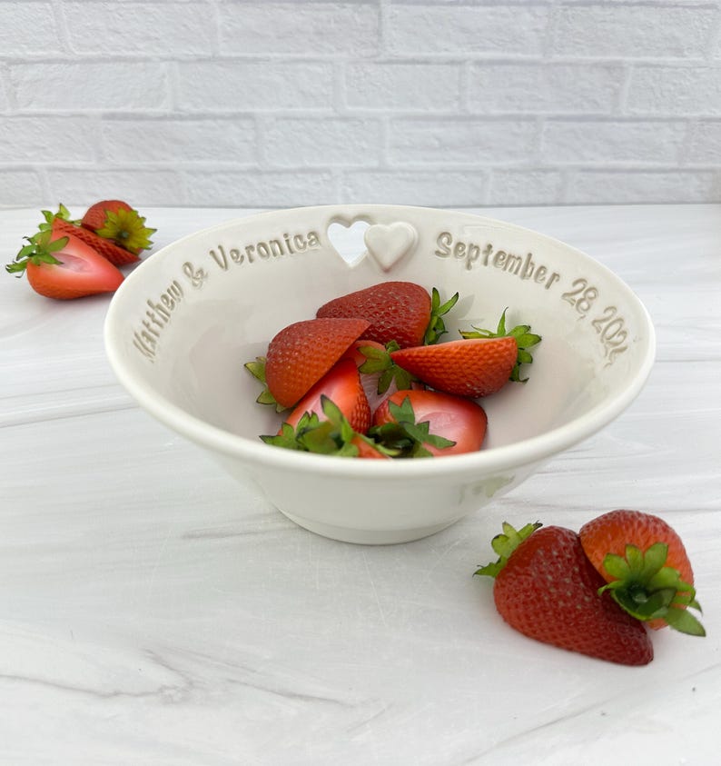 May include: White ceramic bowl with a heart-shaped cutout and the inscription "Matthew & Veronica September 28, 2021". The bowl is filled with fresh strawberries.