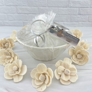 May include: A white ceramic bowl with a gift wrapped in clear cellophane and tied with a silver ribbon. The gift is surrounded by small, white wooden flower decorations. The gift tag reads "The Good Stuff by Katie".