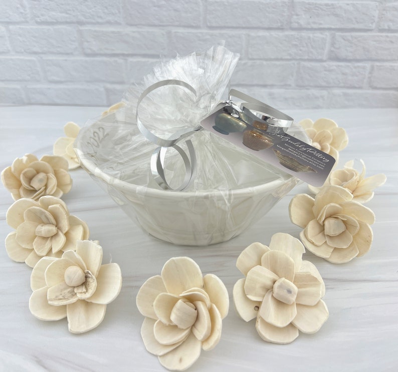 May include: A white ceramic bowl with a gift wrapped in clear cellophane and tied with a silver ribbon. The gift is surrounded by small, white wooden flower decorations. The gift tag reads "The Good Stuff by Katie".