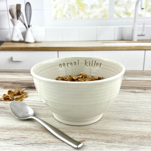May include: White ceramic bowl with the words "cereal killer" inscribed on the side. The bowl is filled with cereal and a silver spoon is on the table next to it.