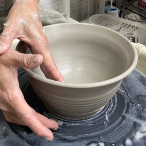 May include: A pottery wheel with a gray ceramic bowl being shaped on it. The bowl is being formed by hand.