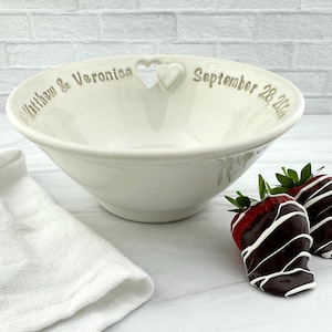 May include: White ceramic bowl with a heart cutout and the text "Matthew & Veronica September 28, 2021". Two chocolate-covered strawberries are on a white surface next to the bowl.