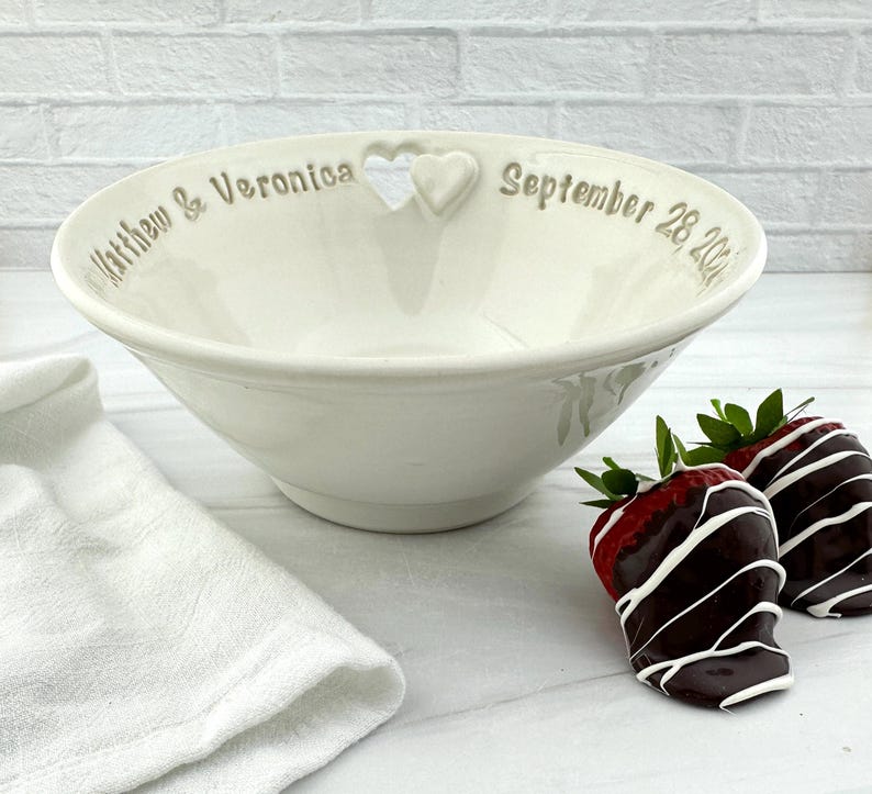 May include: White ceramic bowl with a heart cutout and the text "Matthew & Veronica September 28, 2021". Two chocolate-covered strawberries are on a white surface next to the bowl.