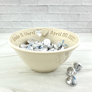 May include: A white ceramic bowl with a heart-shaped cutout and the inscription "Erwin & Merel April 30, 2023". The bowl is filled with silver foil-wrapped chocolates.