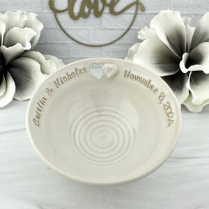 May include: White ceramic ring dish with a heart-shaped cutout and the inscription "Caitlin & Nicholas November 8, 2024".